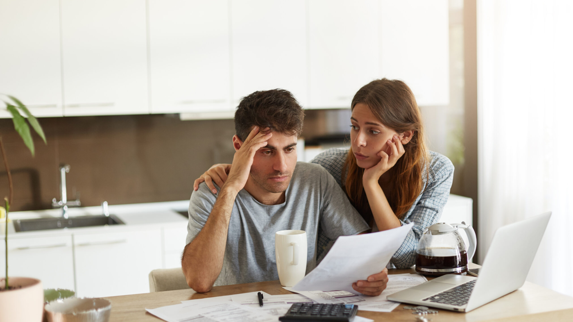 Depressed young Caucasian couple calculating family budget in kitchen, trying to find a way to make both ends meet, having stressed and frustrated looks: caring wife embracing her unhappy husband